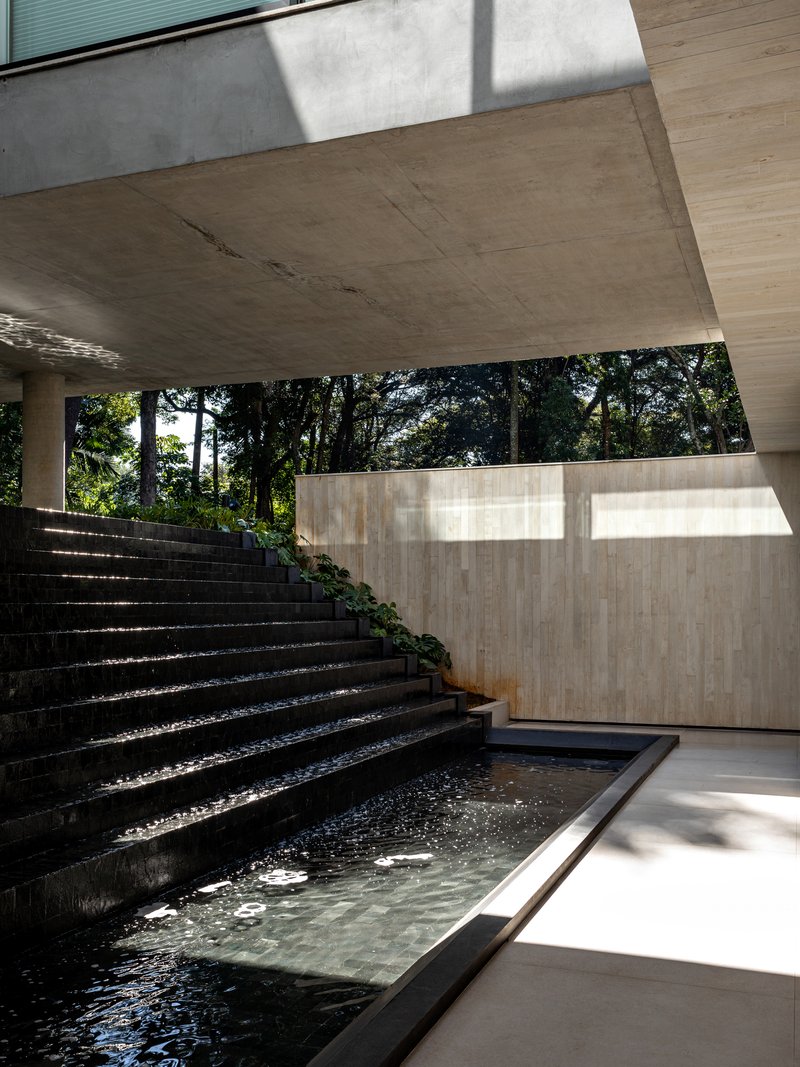 Reflecting pool bordered by stone steps and board-formed concrete walls beneath a deep overhang with filtered light