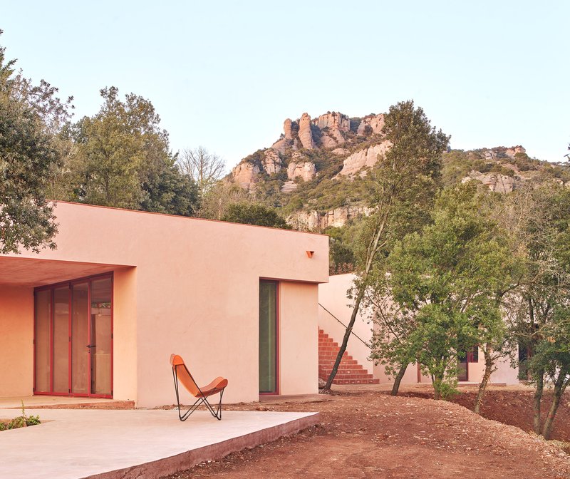Pink stucco facade with timber-framed glazing and an orange butterfly chair beneath rocky hillside vegetation