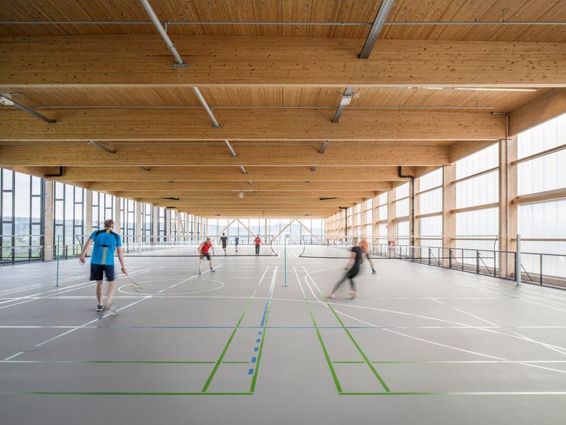 Interior sports hall with exposed timber beams and glazed sidewalls with people playing badminton