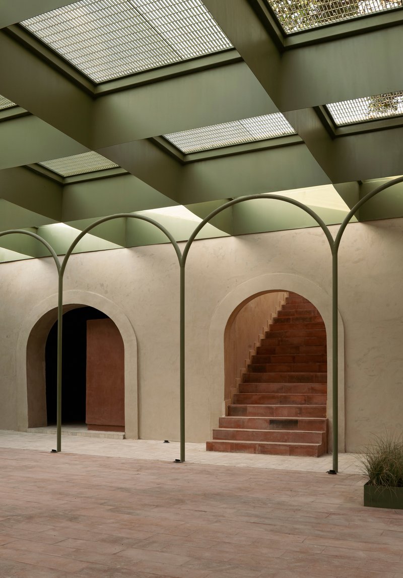 Arched openings framing red terracotta stairs ascending through a cream stone wall beneath a skylit ceiling