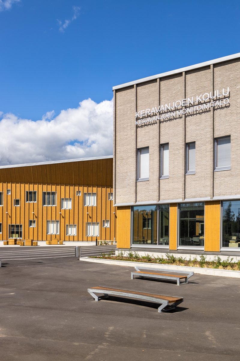 Glass entry vestibule between timber cladding and beige brick facade with wooden benches in foreground