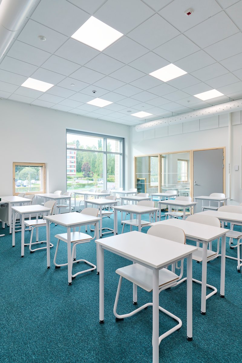 Classroom with white tables on blue carpet showing timber-framed glazed partitions and windows to exterior landscape