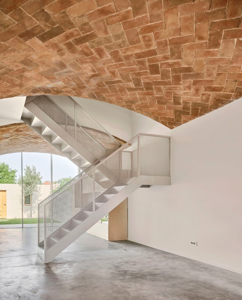 Perforated metal staircase beneath a herringbone brick vault with garden visible through glazed opening