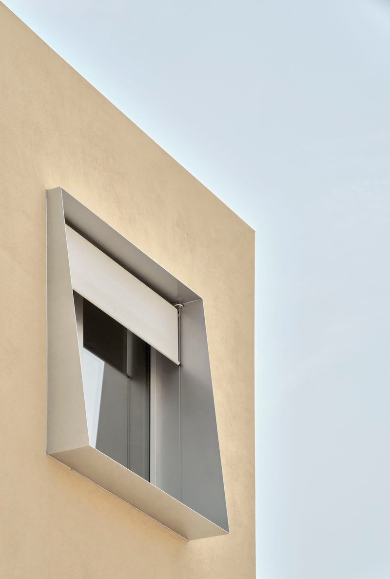 Close-up of recessed window opening with angled metal frame and roller shutter on cream facade