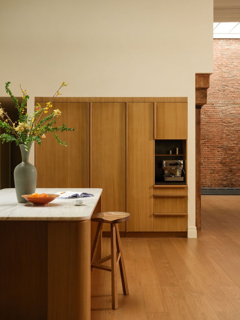 Light timber cabinetry wall with integrated appliances beside an exposed brick wall in afternoon light