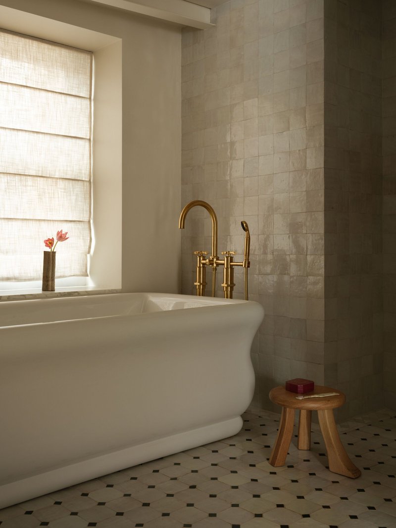 Freestanding white bathtub with brass faucet beside window with woven shade and wooden stool