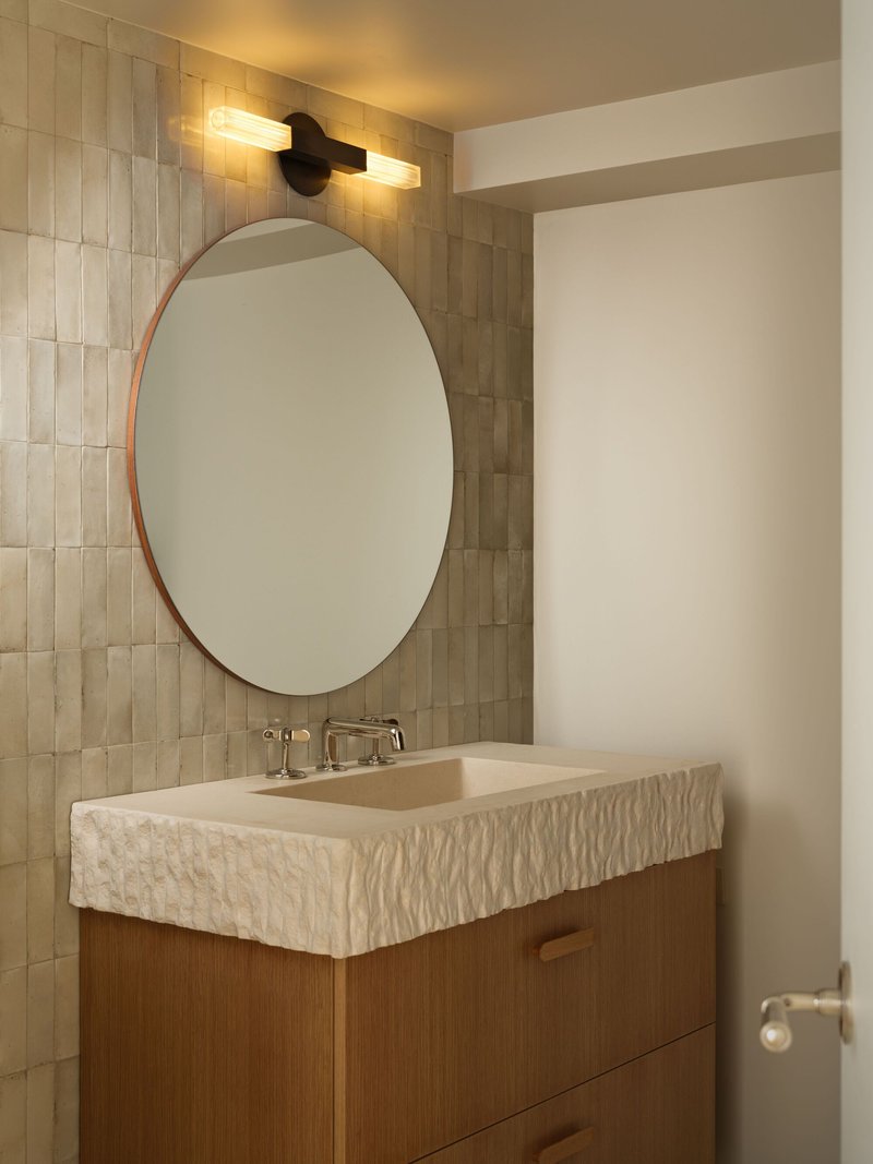 Oval mirror above vanity with textured stone countertop and timber base cabinet under wall sconces