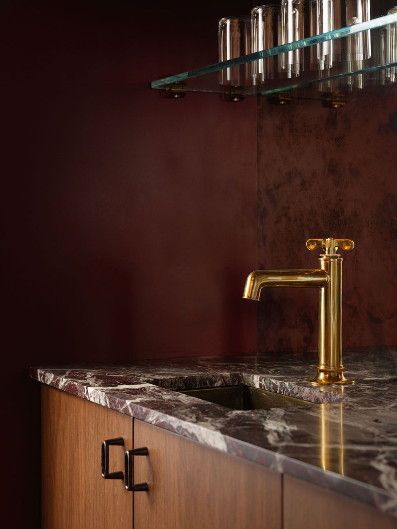 Brass faucet on burgundy veined marble counter against oxidized copper wall panel with glass shelf