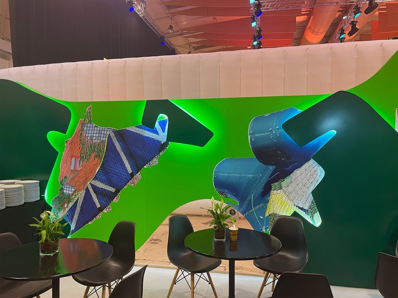 Green booth wall displaying architectural model collages above black chairs and round tables