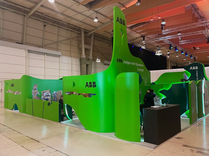 Long view of the exhibition pavilion showing green sculptural partitions and reception counters under exposed structure