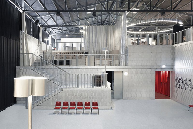 Triple-height interior hall with exposed steel trusses, metal railings, concrete platforms and row of red chairs