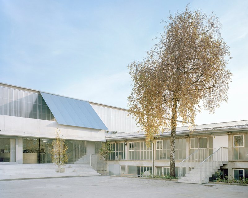 Courtyard view with pale corrugated metal facades and an angular gabled volume beneath a birch tree