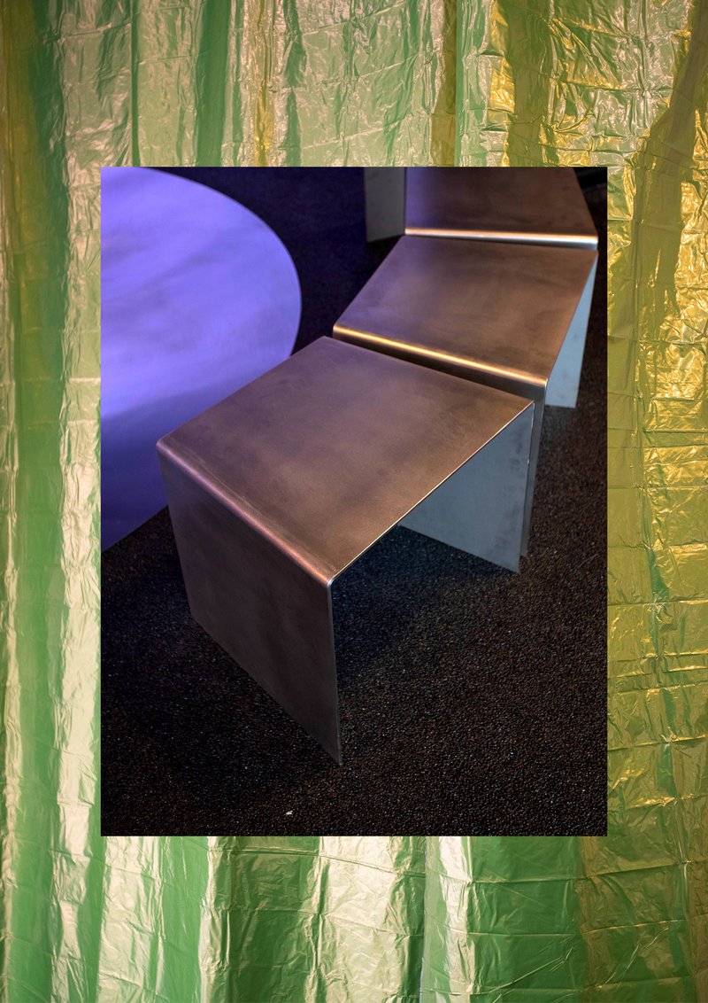 Angular metal bench framed by green and gold foil curtains with a purple circular element visible