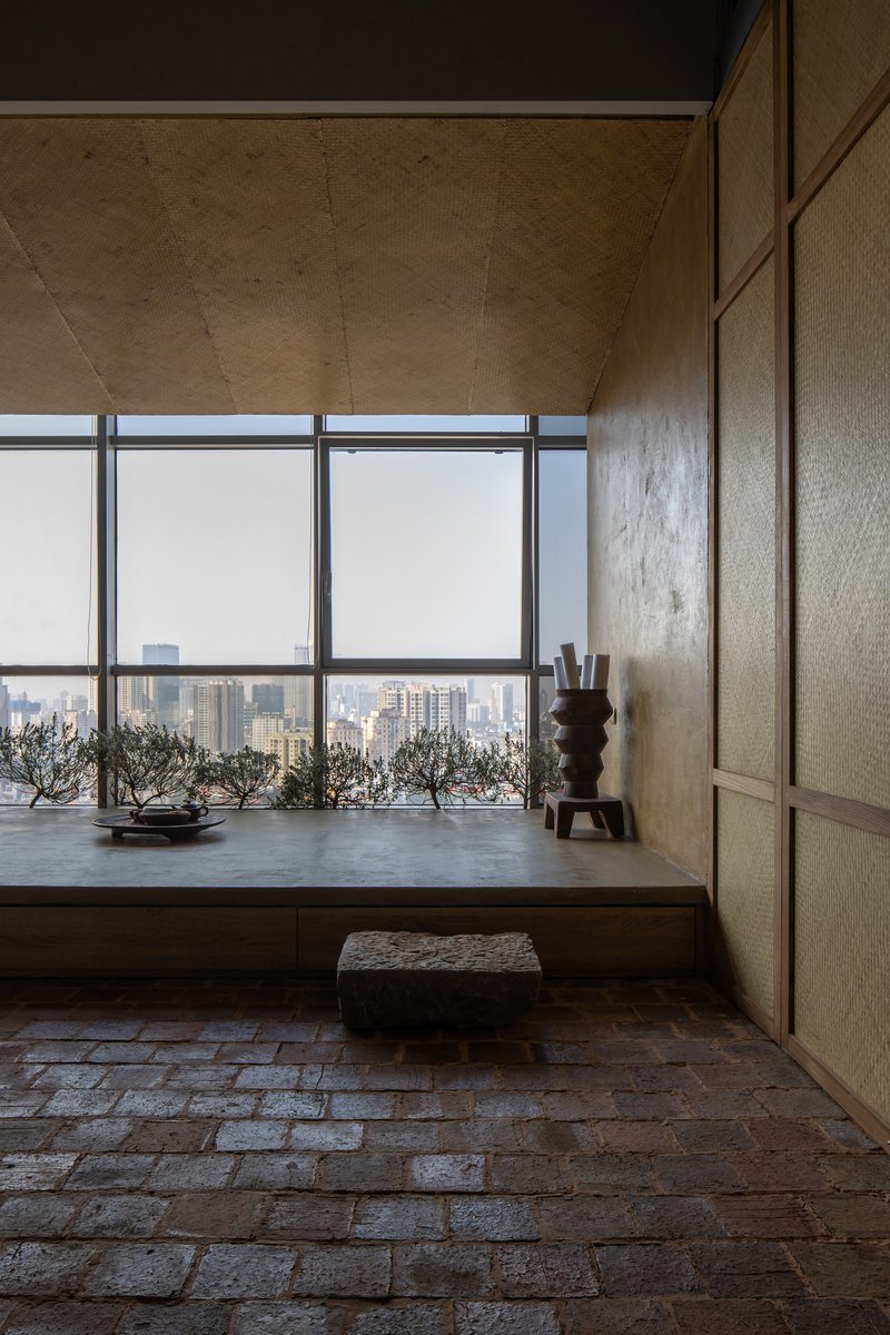 Raised timber platform with potted bonsai trees and floor cushion overlooking hazy cityscape through full-height glazing