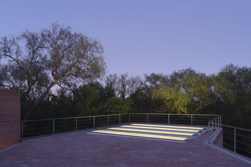 Rooftop terrace with horizontal brick paving, cable railing, and illuminated skylight panel at dusk