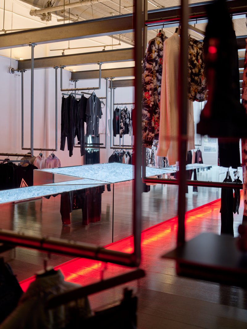 Retail interior with steel clothing racks, marble display tables, and red floor lighting beneath dark wood flooring
