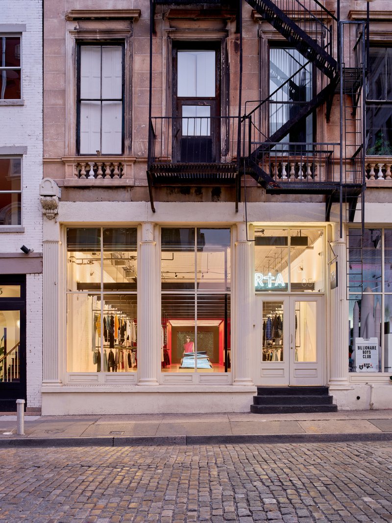 Street facade with white storefront windows below a stone building with black fire escape at dusk