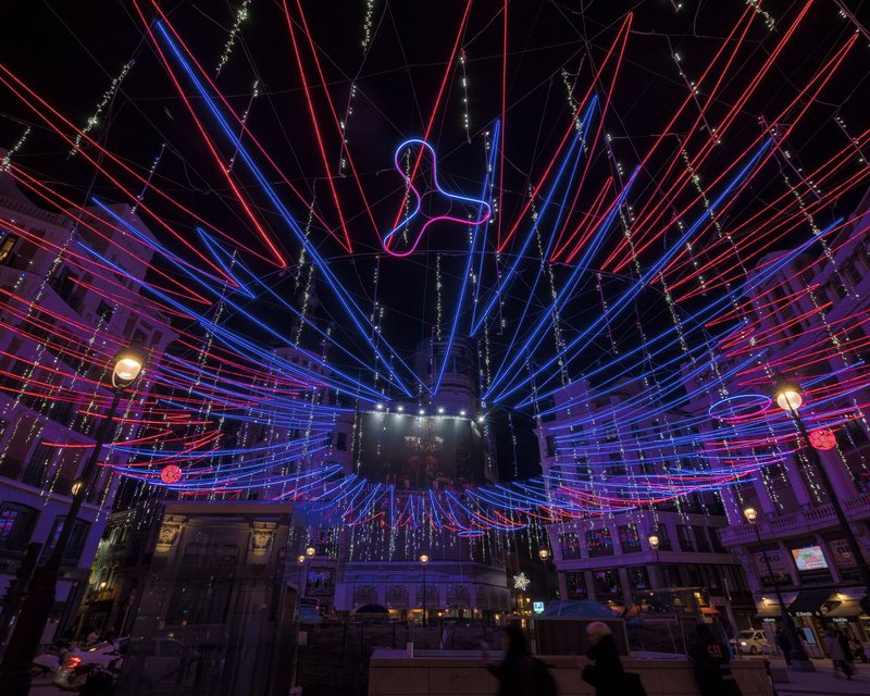 Wide view of LED light installation radiating from central pillar over pedestrians in an urban plaza