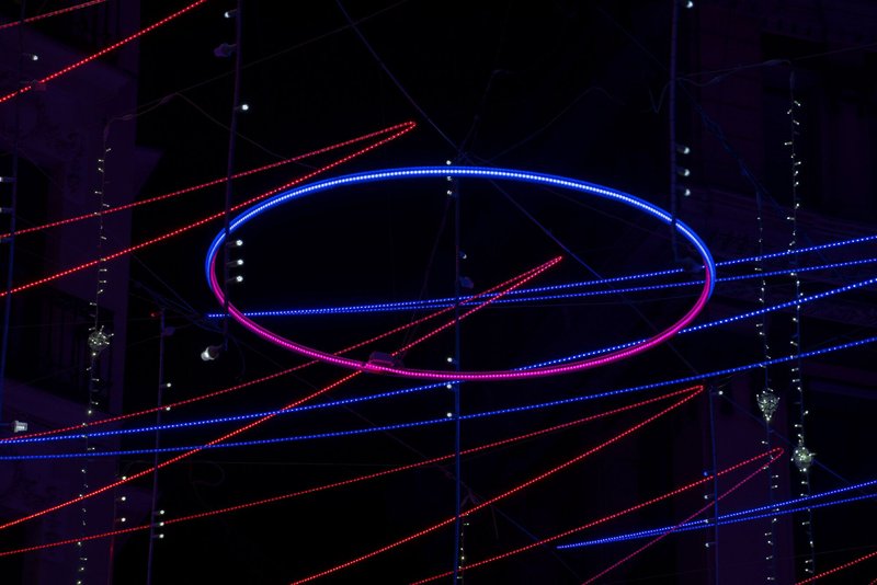 Illuminated elliptical ring with blue and pink LED lights suspended among horizontal light strands at night