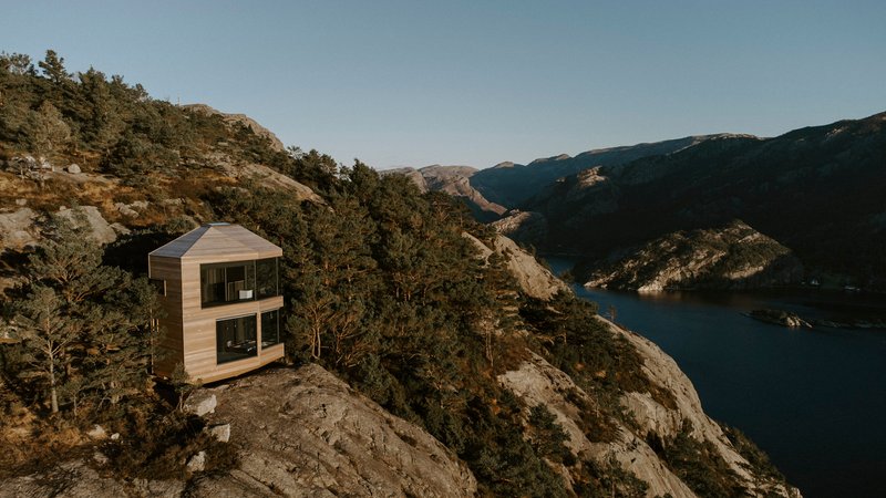 Two-story timber-clad cabin perched on a rocky outcrop above a fjord in late afternoon light