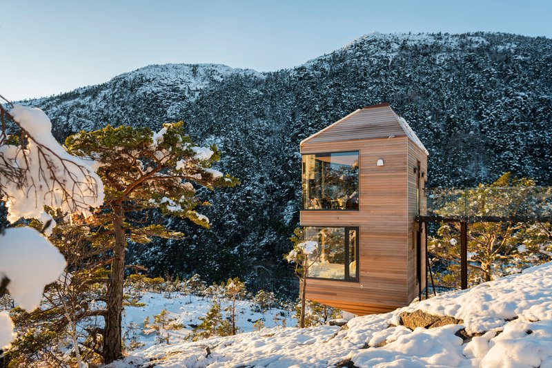 Hexagonal timber-clad structure with floor-to-ceiling glazing set among snowy pines and rock outcrops