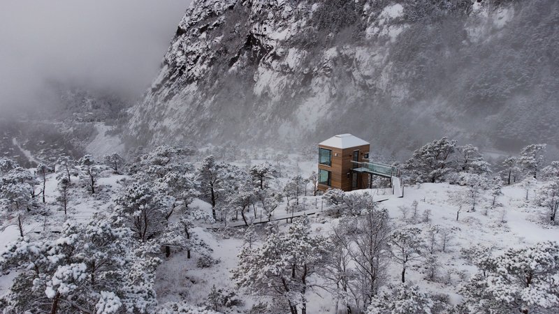 Drone view of the timber cabin in a snowy clearing against a rocky cliff in fog