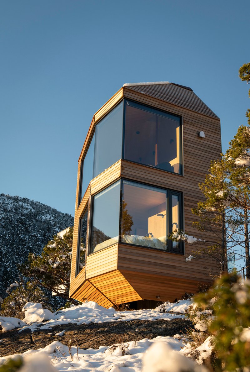 Faceted timber facade with floor-to-ceiling glass panels cantilevering over snowy rock in morning sun