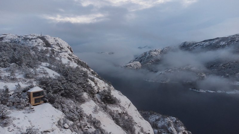 Distant view of the cabin on a snow-dusted slope above a misty fjord