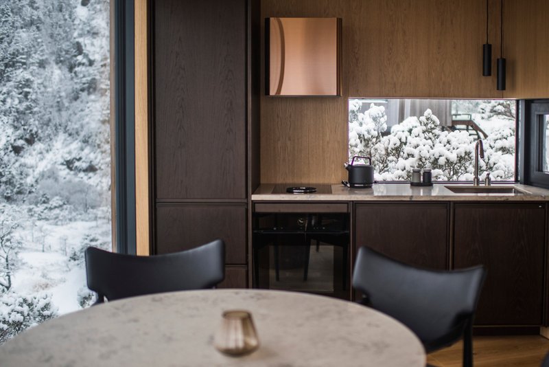 Kitchen with dark wood cabinetry and bronze fixtures framed by floor-to-ceiling windows overlooking snow-covered trees
