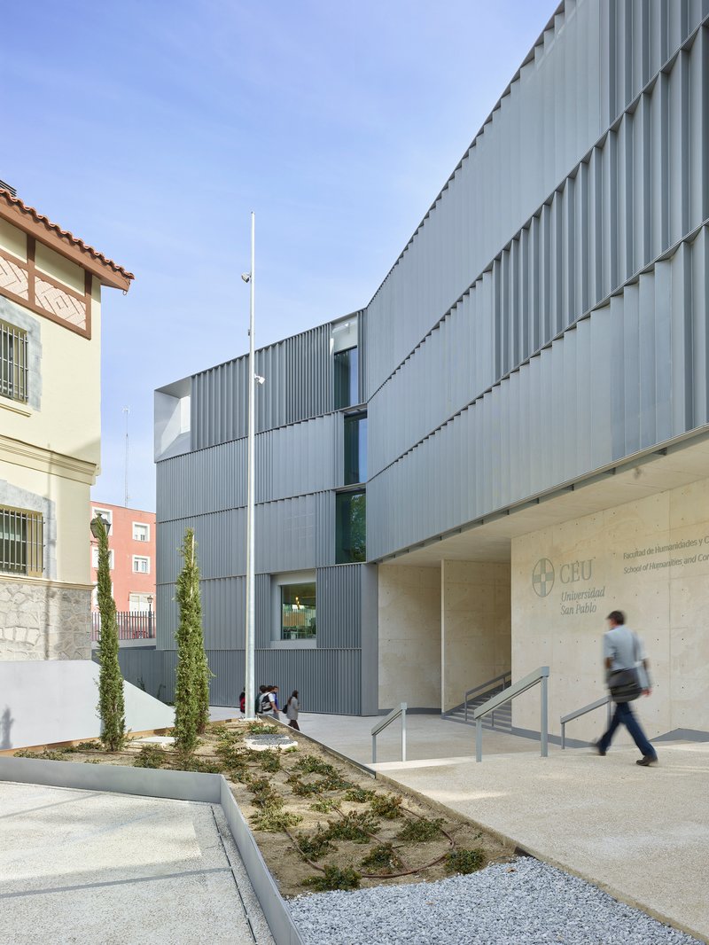 Entrance courtyard with corrugated metal facade, planted beds with cypresses, and a person walking past