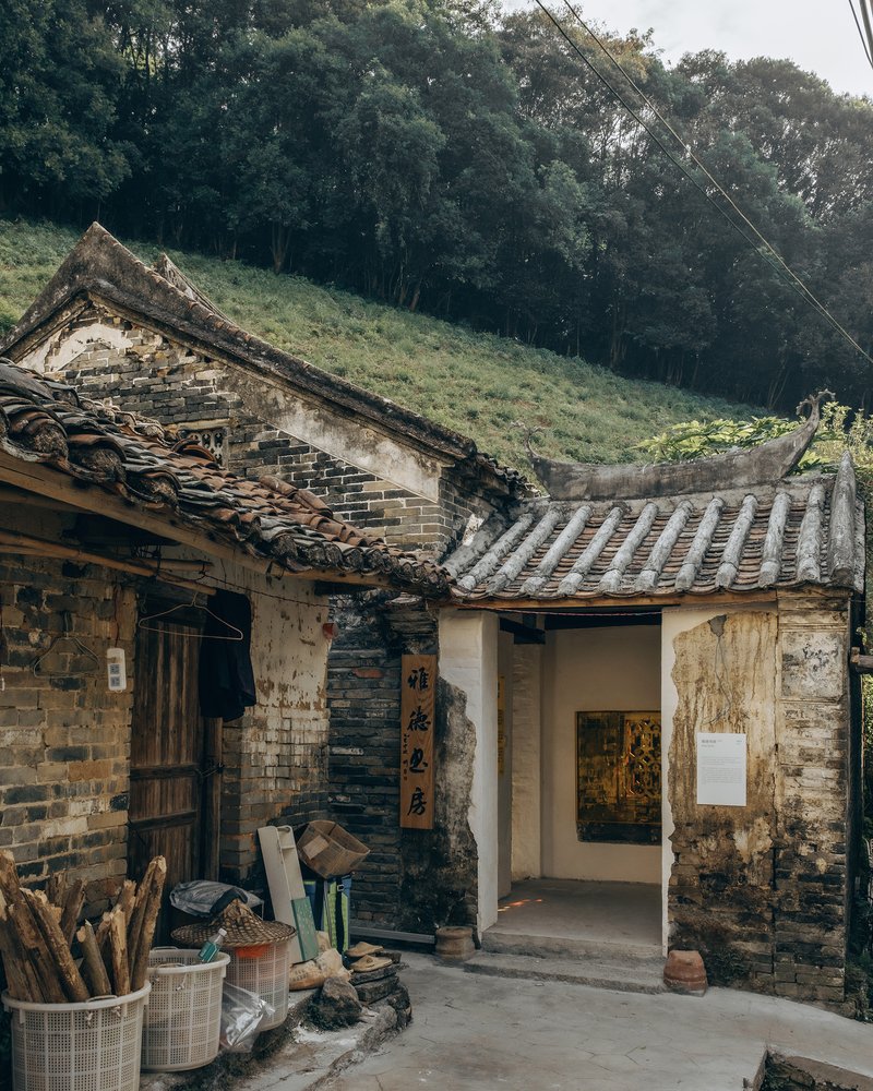 Weathered brick courtyard entrance with curved tile roofs and stacked firewood beneath a forested hillside