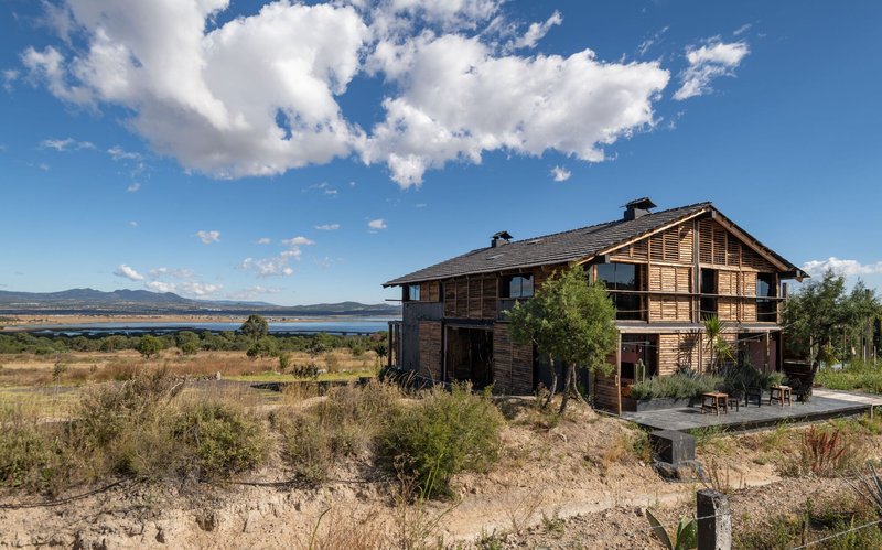 Timber slatted facade with gable roof overlooking native grassland and distant lake under white clouds
