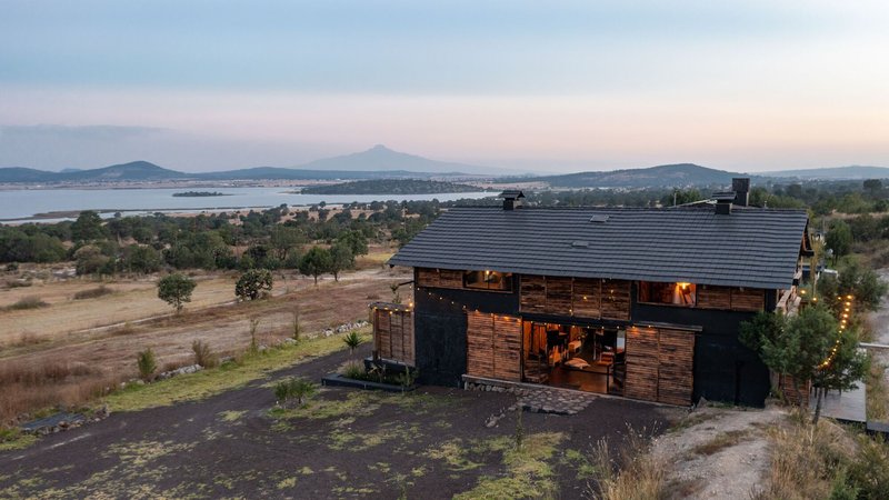 Aerial view of the timber-clad structure with lit interiors at dusk above sparse landscape and distant lake