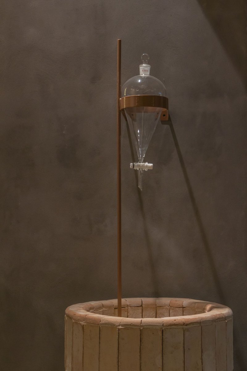 Glass separatory funnel suspended over a circular timber basin on a copper stand against plaster wall