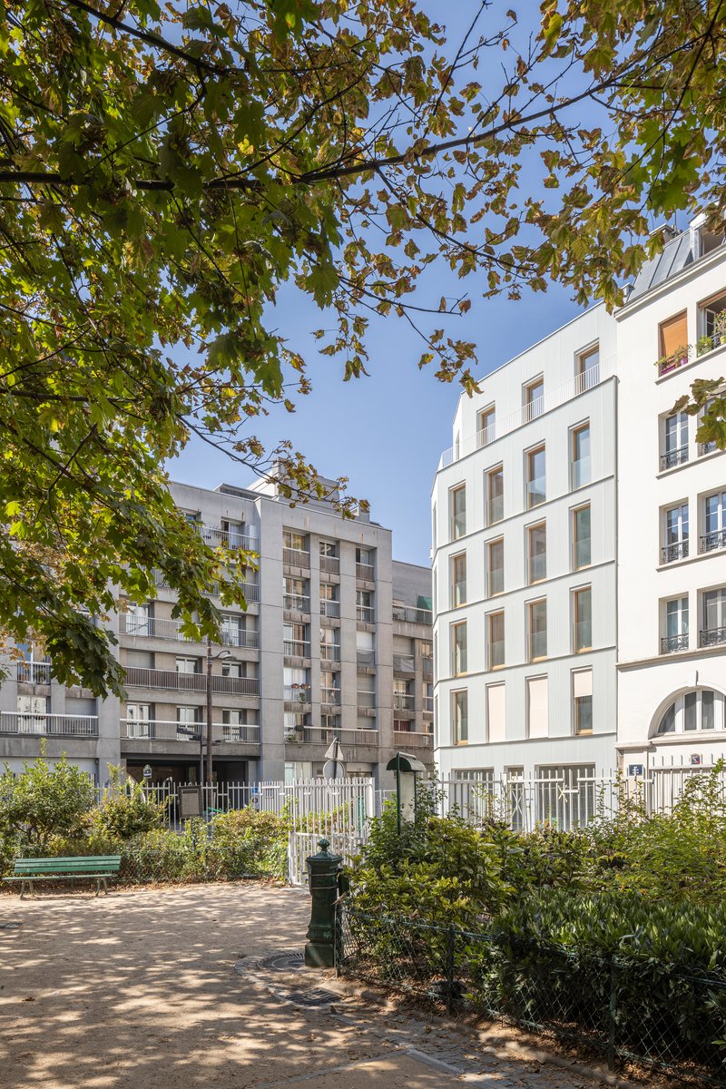 Rear courtyard view of white building through autumn tree canopy with planted beds along metal fence