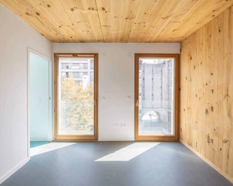 Interior with grey flooring, plywood ceiling and wall, and two windows facing treetops and neighboring buildings