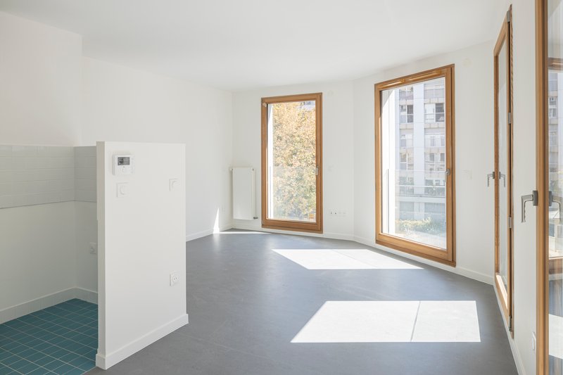 Open plan interior with grey floor, white walls, timber casement windows, and sunlight patches