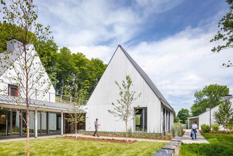 White vertical-board gable building with young trees in a planted lawn under partly cloudy skies