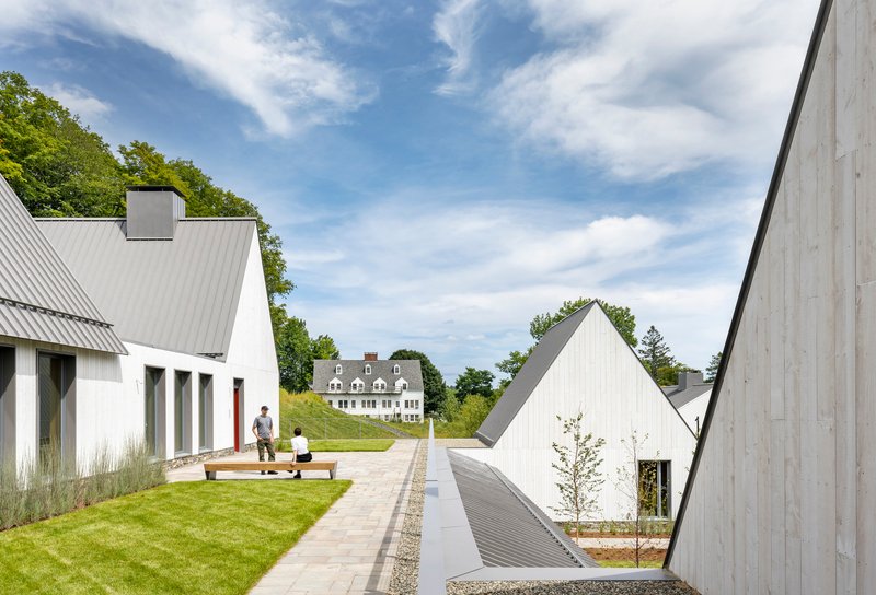Central paved pathway between gabled volumes with two figures on a lawn under scattered clouds
