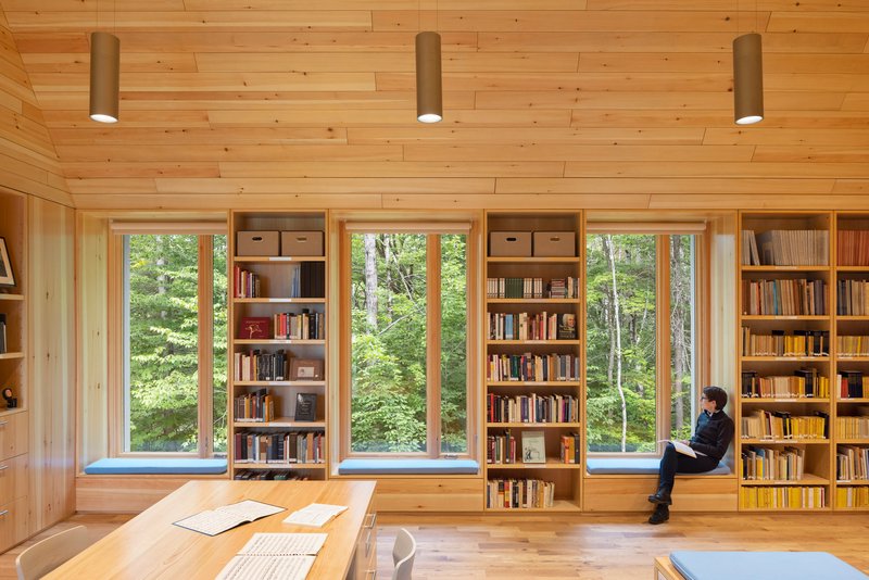 Light timber library with built-in bookshelves and tall windows overlooking summer foliage, person reading in windowsill