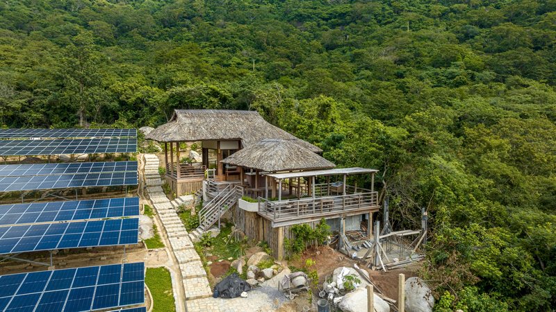 Aerial view of thatched-roof pavilions adjacent to solar panel array amid dense forest