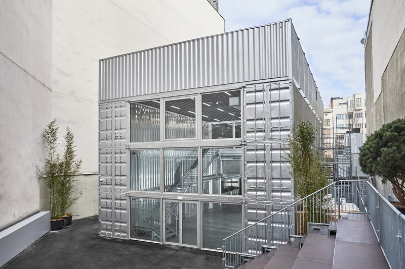 Three-story corrugated metal structure with glass openings and an external metal staircase under daylight