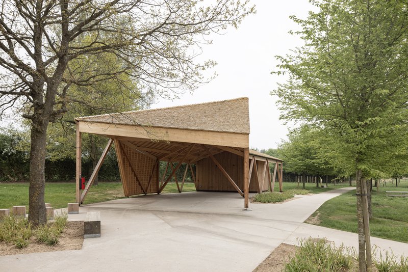 Timber pavilion with diagonal braced columns and woven lattice walls along a concrete pathway in spring