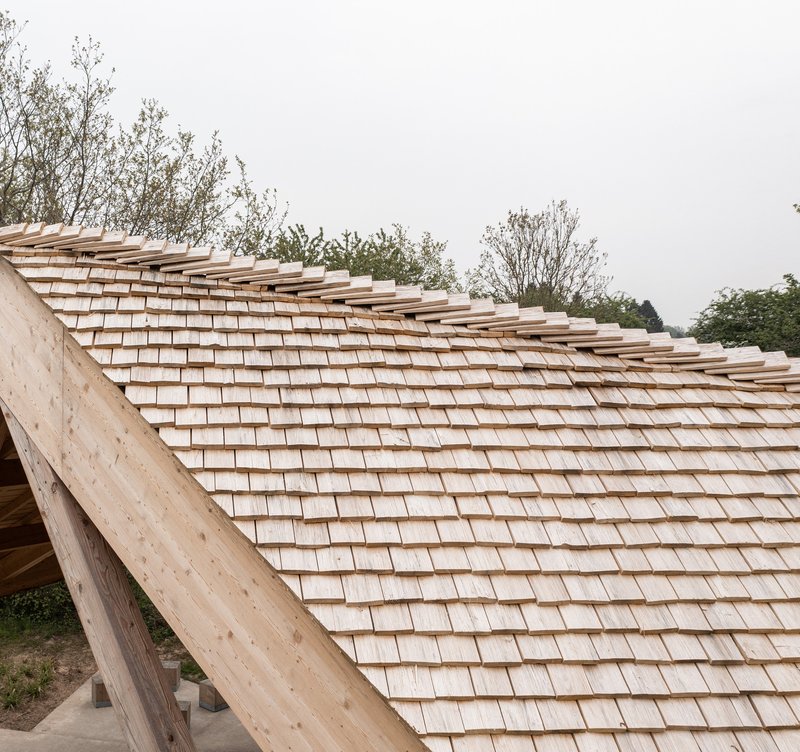 Wood shingle roof with exposed rafter ends curving over a timber structure under overcast skies
