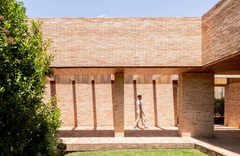 Covered walkway with timber ceiling and brick columns as a figure walks through midday light