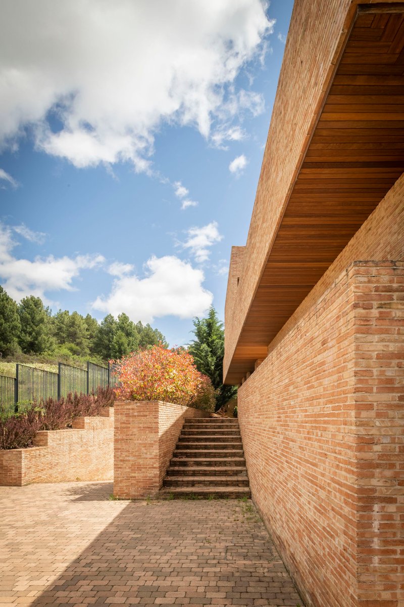 Brick-paved courtyard with stepped planters and timber-lined roof soffit beneath a blue sky with clouds
