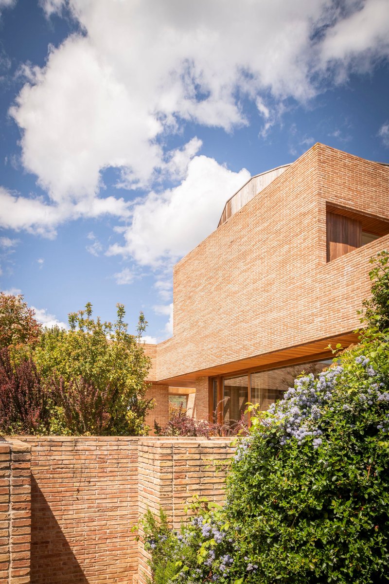 Two-story brick facade with timber soffit and glazed opening above a low retaining wall with flowering shrubs