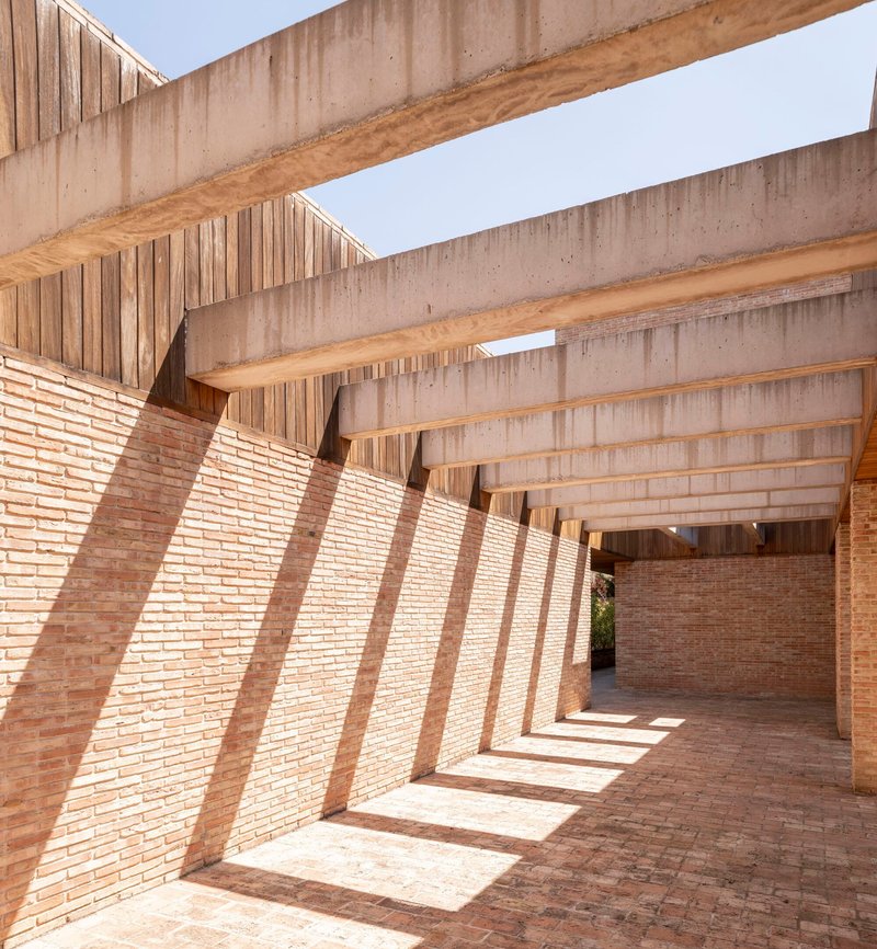 Covered brick walkway with exposed concrete beams casting diagonal shadows across the paved floor