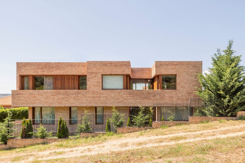 Two-story brick facade with recessed timber-framed windows set above a sloping meadow and wire fence