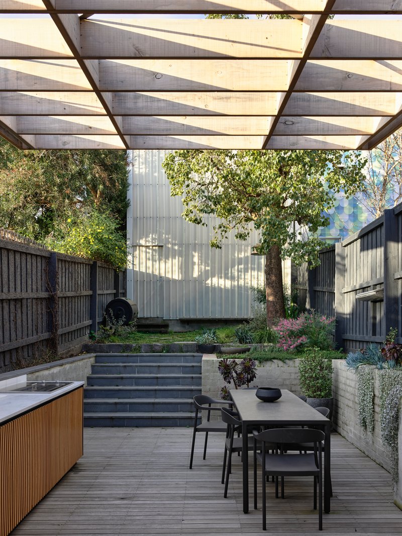 Timber deck with dining table beneath translucent canopy facing concrete steps and planted courtyard